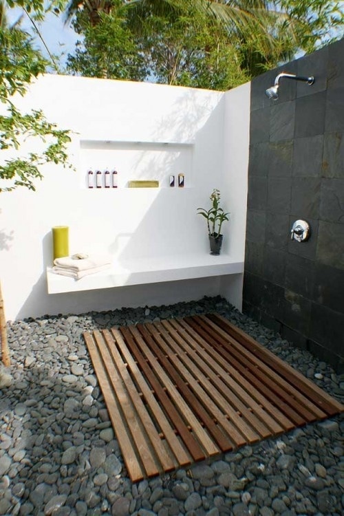 una doccia esterna minimalista con pavimento in pallet, ciottoli, pareti bianche e nere e mensole incorporate