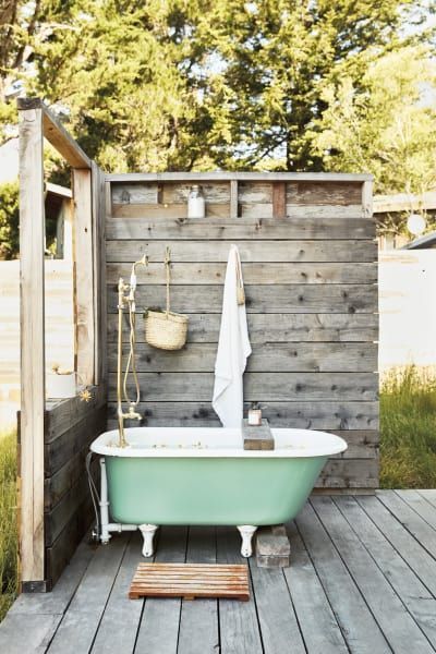 un angolo del bagno rustico all'aperto con schermi in legno stagionato, una vasca verde, un tappeto per pallet e alcuni accessori