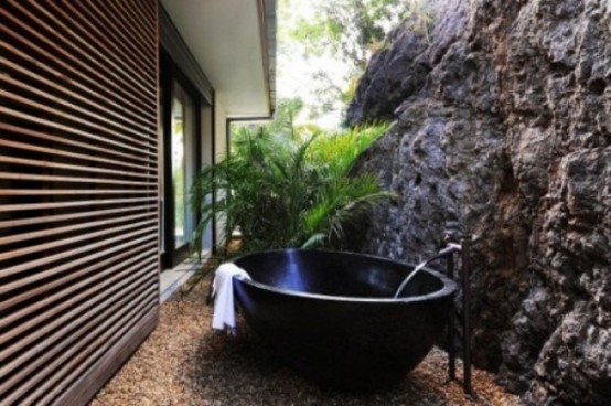 un bagno esterno tropicale con un muro di pietra, una vasca da bagno in metallo e piante in vaso