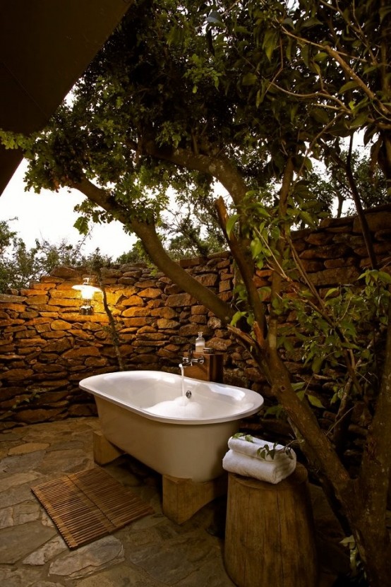 un bagno esterno rustico con un muro in pietra, una vasca su supporti, ceppi come tavolini e alberi viventi