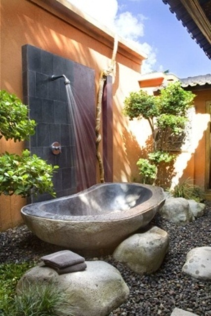 un bagno esterno ispirato alla natura con pietre, una vasca in metallo, ciottoli e vegetazione piantumata