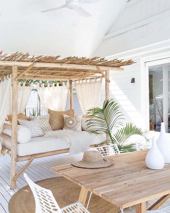 un'accogliente terrazza tropicale con un tavolo in legno, sedie in metallo bianco, una panchina con tetto e tanti cuscini