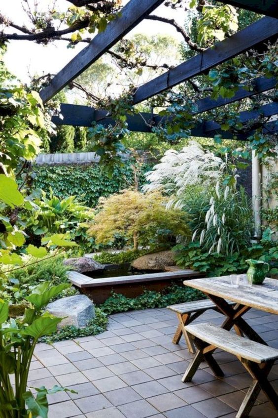 un bellissimo giardino di una casa a schiera con vegetazione piantumata, erbe lussureggianti e materiche, uno stagno, piastrelle e un set da pranzo in legno
